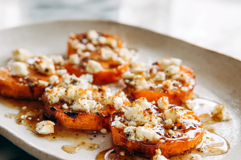 Overhead view of fresh ingredients on a wooden cutting board: two large orange sweet potatoes, a small bowl of crumbled white goat cheese, a jar of golden honey, fresh thyme sprigs, olive oil in a glass bottle, and small bowls of sea salt and black pepper