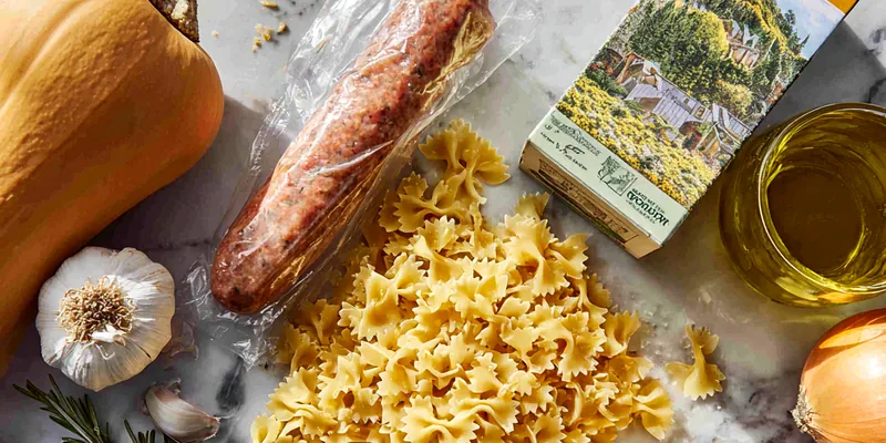Overhead view of colorful Italian sausage and vegetable pasta in a black ceramic bowl on marble surface, showing golden bow tie pasta mixed with browned sausage slices, vibrant red and yellow bell peppers, green zucchini, and roasted Brussels sprouts, garnished with fresh parsley and grated Parmesan cheese
