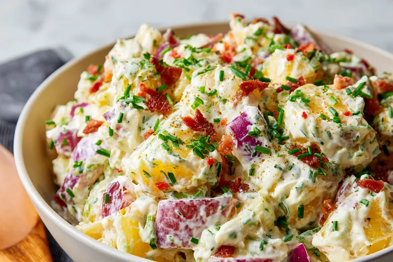 Flat lay of potato salad ingredients on marble counter including red potatoes, bacon, eggs, mayo, sour cream, chives and spices
